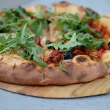 Pizza z rukolą, oliwkami i papryką na twardym cieście do pizzy, podana na drewnianej desce.