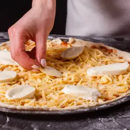 Ręka układa kawałki sera na pizzy, która już ma starty żółty ser. Jaki ser do pizzy? Mozzarella i żółty ser to świetne połączenie!